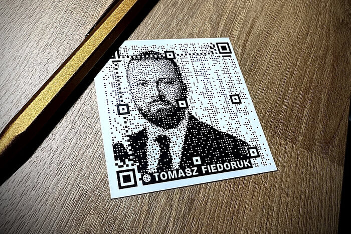 A printed business card with a portrait formed by QR dots lies on a table beside a pen.